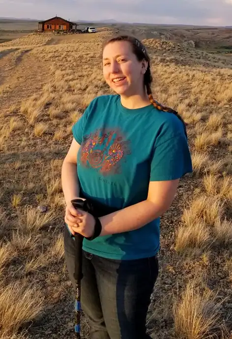 A woman stands smiling on a dirt trail surrounded by golden prairie grass under a wide, open sky. She wears a teal shirt with a colorful design and holds a walking stick. In the distance, a cabin sits on the horizon, and a small white dog stands on the trail. A wooden sign labeled “HORN” with a painted landscape detail is partially visible in the foreground on the right. The scene is bathed in warm evening light.