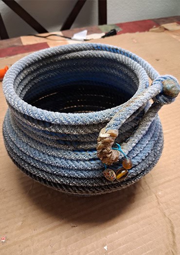 A round, deep basket crafted from blue and gray rope with a knotted loop and bead embellishment near the rim. The piece is displayed on a cardboard-covered table, suggesting a work-in-progress or artist’s studio setting.