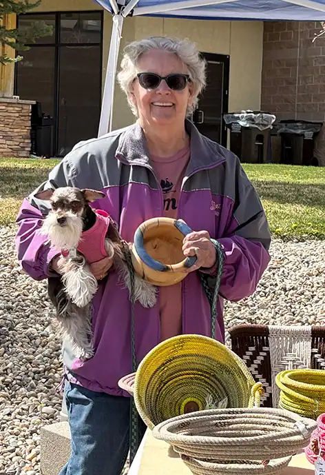 Smiling woman wearing sunglasses and a purple jacket, holding a small black and white dog in a pink vest and a handwoven bowl, standing behind a table displaying colorful coiled rope baskets at an outdoor market