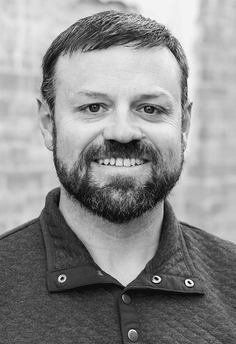 A man with short dark hair and a beard smiles at the camera, wearing a quilted button-up pullover against a blurred brick wall background.