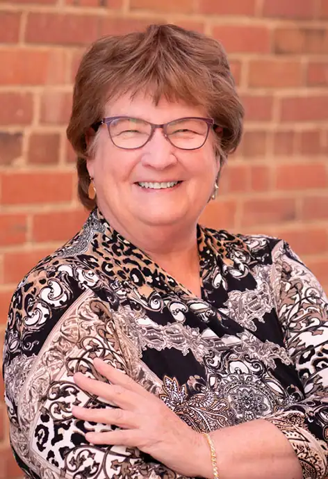 Professional headshot of Lynn Jones, a woman with short gray hair, glasses, and a multi-colored scarf, smiling gently in front of a light-colored indoor wall