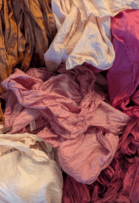 A close-up of naturally dyed silk scarves in soft, earthy tones including shades of rose, peach, ivory, and ochre, showing their texture and organic folds.