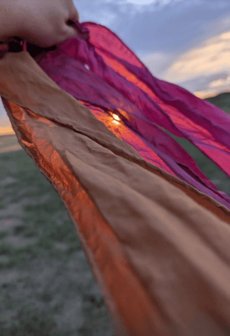 Hand-dyed silk scarves in warm sunset colors—rust, pink, and burgundy—flow in the breeze, held up against a dramatic prairie sky at dusk.