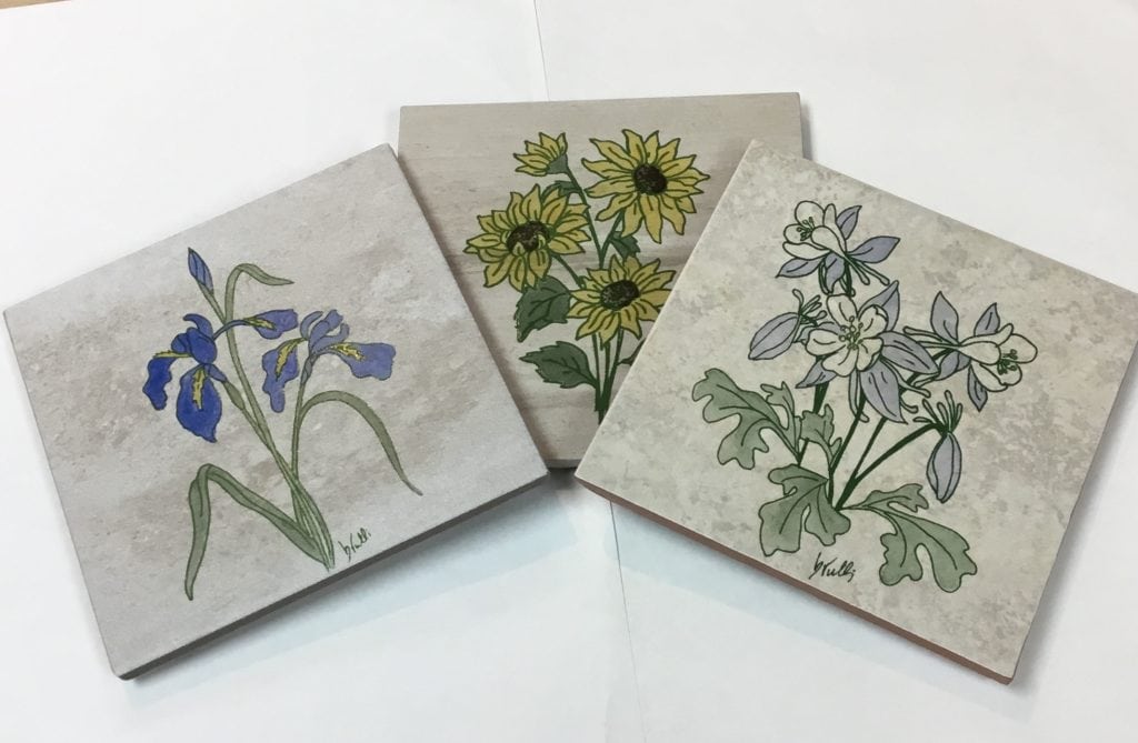 Three square ceramic tiles laid out on a white background, each painted with a different native Wyoming wildflower—blue iris, yellow sunflowers, and pale blue columbine—on a natural stone-textured surface.