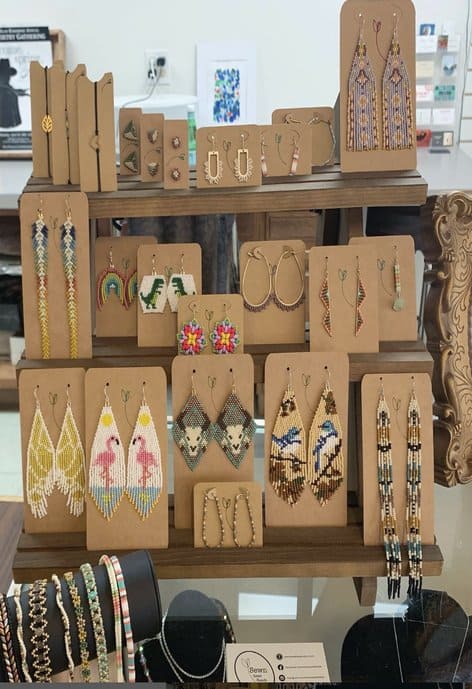 A display rack of colorful beaded earrings in various designs including geometric patterns, rainbows, flamingos, birds, and florals—each set mounted on brown backing cards with a heart leaf logo.