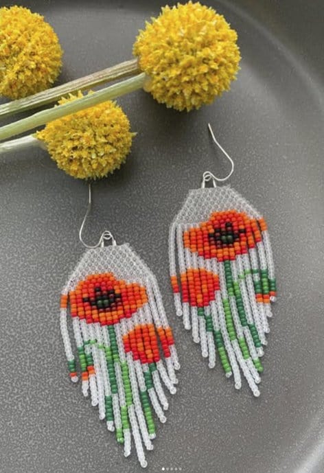 Hand-sewn beaded earrings with bright red and orange poppy flower designs and white fringe, displayed next to yellow pom-pom flowers on a dark textured background.