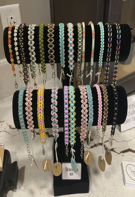 A black jewelry stand holding two rows of hand-sewn beaded bracelets in bright pastel colors, metallics, and floral patterns, labeled “Bracelets $25” with price tags attached.
