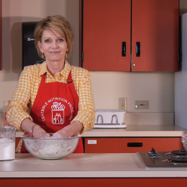 woman in kitchen with CNP apron smiling while hand mixing recipe