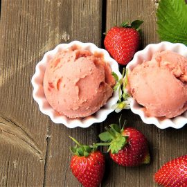 Pink dessert in 3 white bowls, surrounded by fresh strawberries on a wood background.