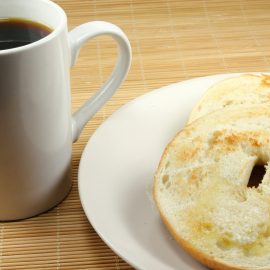 toasted sliced bagel on white plate next to white mug with black coffee; both on wooden table