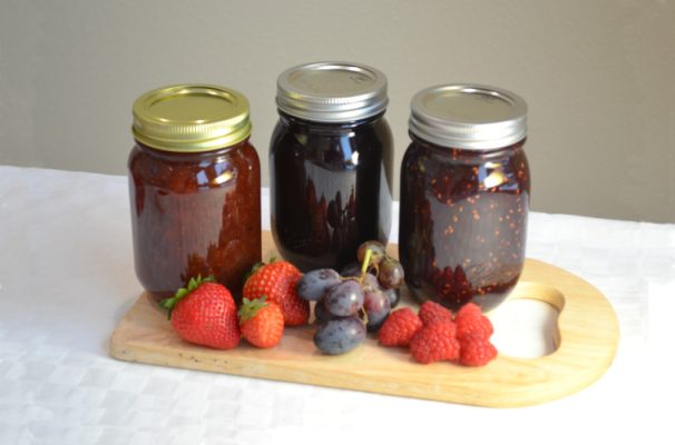 Three jars of jam with strawberries and grapes on a wooden board