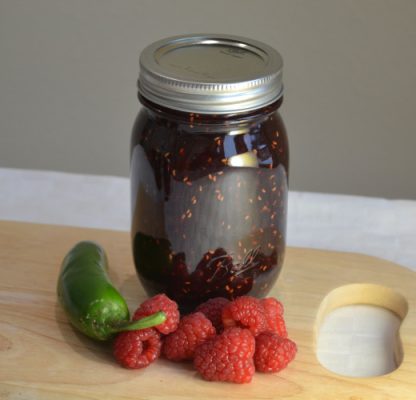 Jar of hot sauce with a jalapeno pepper and raspberries on a wooden cutting board.
